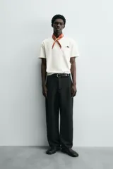 Camiseta de corte relajado (relaxed fit) color blanco hueso, con cuello redondo, manga corta y bolsillo plastrón en el pecho. Presenta un bordado pequeño y contrastante de una palmera en el área del bolsillo.