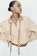 Camisa de popelín color beige con cuello solapa y lazada frontal en contraste con estampado de rayas marrones. Presenta mangas largas abullonadas con puños elásticos, bolsillos con solapa en el pecho y bajo elástico.
