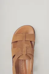 Sandalias planas de gamuza color marrón, con diseño de tiras cruzadas en el empeine y cierre con hebilla metálica dorada en el tobillo. Cuenta con plantilla de cuero y suela de goma con textura.