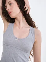 Musculosa de punto pointelle color gris melange, con escote redondo, detalle de picot en los bordes y una pequeña moña decorativa en el centro del pecho.