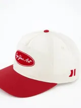 Gorra de béisbol color crema con visera y botón superior rojos. Presenta un logo bordado en el frente con la frase "Sign Your Art" en rojo.