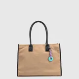 Tote bag de gamuza en color beige con detalles en negro, incluyendo asas, bordes y refuerzos. Cuenta con un charm colgante decorativo en tonos violeta y turquesa.