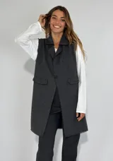 Chaleco largo de paño color gris oscuro, con solapas, un botón frontal y dos bolsillos de ojal. Presenta costuras decorativas en contraste y bordes deshilachados.