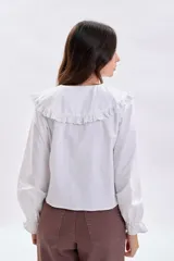 Camisa blanca de manga larga con cuello grande estilo babero con detalles de bordado inglés (broderie), cierre frontal con botones y puños elásticos con volados.