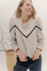 Camisa de lino color beige con mangas abullonadas y detalles de puntilla negra en forma de V en el frente. Presenta cuello a la base con borde desflecado, cierre frontal con botones forrados y puños con alforzas.