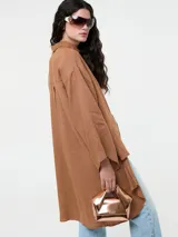 Camisa larga de lino color marrón, con corte oversize, mangas anchas y bolsillo frontal de cuero sintético en tono contrastante. Presenta cierre frontal con botones y diseño de bajo irregular.