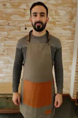Delantal unisex de lona color gris con bolsillo de cuero marrón, tiras de cuero ajustables con hebillas metálicas y logo grabado en la parte inferior. Confección artesanal.