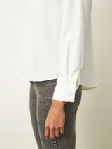 Blusa de color blanco con cuello en V y mangas largas con puños abotonados. Presenta un diseño de corte recto y holgado.