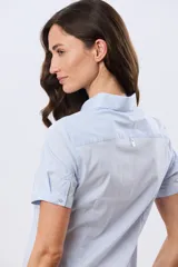 Camisa celeste rayada de manga corta, con cuello camisero y cierre frontal con botones. Presenta un corte entallado y un pequeño detalle de placa metálica en el borde inferior.