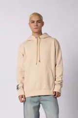 Buzo tipo canguro color beige con capucha y cordones ajustables. Presenta un bolsillo frontal tipo canguro y un parche decorativo en la manga derecha.