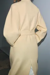 Abrigo largo color crema confeccionado en mezcla de lana. Diseño oversize con cuello en V, mangas largas y cinturón ajustable con lazada.