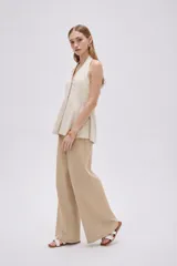 Blusa de algodón color beige con escote en V, cuello halter y cierre frontal con botones. Presenta un diseño ajustado en el torso con pinzas y pliegues en la parte inferior que generan una caída ligeramente acampanada.