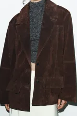 Chaqueta tipo blazer oversize de piel de ante color marrón oscuro, con cuello solapa de muescas, manga larga y bolsillos delanteros con solapa. Cierre frontal con botones.