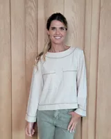 Remera de manga larga en color beige, confeccionada en tejido de punto con costuras a la vista en tono contrastante. Presenta cuello redondo y dos bolsillos frontales tipo parche.