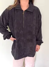 Buzo negro de pana con cierre metálico en el cuello, corte holgado y mangas largas con puños elásticos.