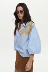 Camisa de algodón a rayas celestes y blancas, con mangas abullonadas y gran estampado floral en el frente.