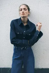 Jeans de tiro alto, pierna ancha y corte recto, confeccionado en denim de algodón rígido de color azul medio. Presenta un detalle de herrajes metálicos en la cintura.