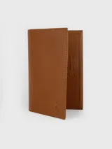 Porta pasaporte de cuero color marrón tostado, de diseño vertical y minimalista con costuras pespunteadas a contratono. Presenta el logotipo de la marca grabado en la esquina inferior y sello interior con la leyenda de autenticidad.