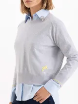 Sweater de tejido de punto en color gris, con cuello redondo, mangas largas y detalle de bordado amarillo en el puño y en el lateral inferior. Presenta un corte recto y estructura ligera.
