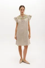 Vestido corto color beige de lino, corte recto, cuello redondo con volados y abertura en forma de gota con lazo. Mangas cortas con volados.