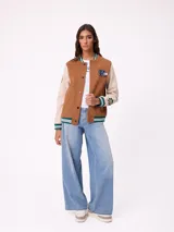 Campera estilo varsity con cuerpo en color marrón y mangas en tono beige. Presenta cierre frontal con botones a presión, cuello, puños y ruedo con franjas tejidas en verde y blanco. Incluye un parche con la letra R en el pecho y detalles bordados en las mangas.