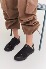 Zapatillas urbanas de color negro con diseño slip-on, caracterizadas por bandas elásticas cruzadas en el empeine y detalles en charol. Cuentan con suela plana de goma al tono.