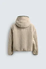 Cazadora de corte relajado confeccionada en tejido de textura borreguillo color beige. Presenta cuello subido con capucha ajustable, cierre frontal de cremallera en contraste verde, mangas largas y bolsillos laterales. El interior está forrado en tejido técnico.