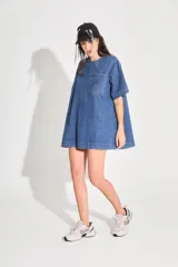 Vestido corto de denim azul con corte holgado, mangas cortas y bolsillo frontal con logo bordado. Cuenta con cierre metálico en la espalda.