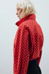 Chaqueta roja con estampado de lunares blancos, cuello subido, manga larga con puños plisados, bolsillos delanteros, bajo ajustable con cordón y cierre frontal con cremallera.