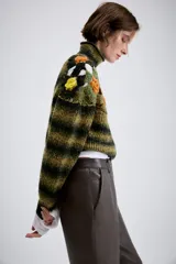 Jersey de punto con cuello subido y manga larga. Presenta un diseño de rayas horizontales en tonos verde y negro, con un panel superior decorado con calados y aplicaciones de flores tejidas en crochet de colores contrastantes.