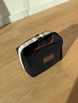 Neceser de neopreno color borgoña, con cierre superior y forro interior. Tiene un asa de mano con una franja azul y blanca.