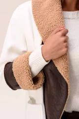 Saco de corte largo en color blanco, con solapas amplias y puños decorados con textura de corderito en color tostado. Presenta detalles en cuero sintético marrón en la parte frontal y en los puños, además de bolsillos laterales.
