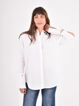 Camisa blanca de corte clásico con cuello camisero y puños anchos destacados. Presenta cierre frontal oculto por tapeta y dobladillo redondeado.