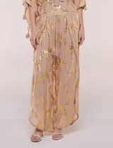 Pantalón palazzo confeccionado en gasa transparente color rosa con diseño estampado metalizado dorado. Presenta cintura elástica con cordón ajustable con borlas, piernas anchas y diseño cruzado al frente con aberturas laterales y detalles de puntillas.