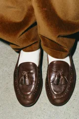 Mocasín de cuero color marrón con detalle de borlas en el empeine y perforaciones decorativas tipo brogue en la puntera y el talón.