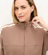 Campera deportiva oversized de microfibra color marrón, con cuello alto, cierre frontal y bolsillos laterales. Presenta bandas laterales contrastantes en las mangas y cordón ajustable en el dobladillo.