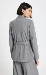 Blazer gris de sastrería con diseño entallado, cuello con solapas clásicas, hombros estructurados y mangas largas. Presenta bolsillos delanteros con solapa y un cinturón a juego con hebilla metálica para ajustar la cintura.