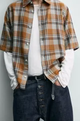 Camisa de corte boxy fit confeccionada en tejido de algodón, con estampado de cuadros en tonos tierra. Presenta cuello solapa, manga corta y cierre frontal de botones.