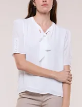Blusa blanca de corte recto y tejido liviano de gasa con forro interno delantero. Presenta escote en V con cuello redondo y lazo central para anudar, mangas cortas con hilera de botones forrados en los laterales y ruedo ligeramente redondeado.