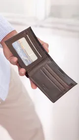 Billetera de cuero marrón oscuro, estilo clásico con doblez. Presenta un bolsillo exterior con cierre metálico en la parte frontal y un compartimento con cierre en el interior posterior. El interior cuenta con un visor para identificación y múltiples ranuras para tarjetas.