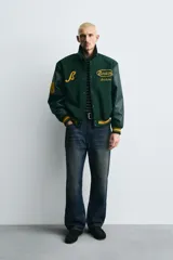Cazadora estilo varsity con cuerpo de mezcla de lana en color verde oscuro y mangas de efecto piel a tono. Presenta cuello solapa, cierre frontal con botones a presión, bolsillos laterales de vivo y puños y bajo acanalados con rayas amarillas. Incluye bordados y parches decorativos en el frente con la inscripción Boston Archive.