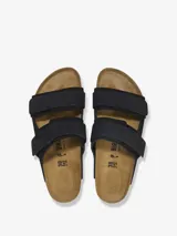 Sandalias Birkenstock modelo Uji de color negro, con dos tiras anchas ajustables mediante cierres de contacto y detalles de remaches metálicos. Confeccionadas con una combinación de gamuza y cuero nobuk, presentan la clásica plantilla anatómica de corcho y suela de goma negra.