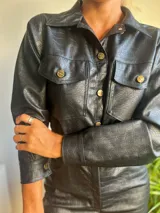 Mono corto negro de cuero sintético con textura croco, cuello camisero, cierre frontal con botones dorados, mangas largas con puños abotonados, bolsillos con solapa en el pecho y aberturas laterales en la cintura.