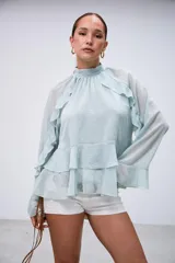 Blusa de gasa color verde agua, con cuello alto fruncido, mangas largas acampanadas y diseño con volados en el frente y hombros.