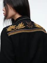 Camisa de color crudo con detalles de bordados en tonos mostaza y marrón en el cuello, hombros, bolsillos frontales y puños. Presenta cierre frontal con botones y dos bolsillos tipo cargo en el pecho.