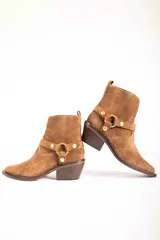 Bota corta de gamuza color marrón, con taco cuadrado y punta fina. Presenta una correa con tachas doradas y una argolla metálica decorativa alrededor del tobillo. Cuenta con cierre lateral dorado.