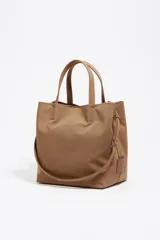 Bolso grande Bimba y Lola color marrón, con exterior de gabardina y detalles en cuero vacuno. Presenta doble asa de mano, correa ajustable, cierre con botón a presión, logo metálico de la marca en dorado y bolsillo interior con cremallera.