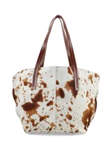 Bolso tipo tote de piel de vaca, con estampado moteado en blanco y marrón. Presenta asas de cuero marrón oscuro y costuras decorativas en forma de X en el centro frontal.