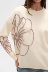Sweater de tejido de punto color crema, con diseño holgado, cuello redondo a la base y mangas largas. Presenta apliques bordados en relieve con forma de flores en tonos rosados en el frente y las mangas.