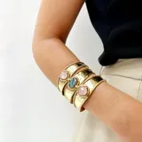 Brazalete ancho de bronce dorado, con textura martillada, compuesto por tres bandas unidas y adornado con tres piedras engarzadas: una central ovalada de nácar blanco y dos laterales cuadradas de nácar blanco.